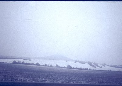 Vintage black-and-white landscape showing snow-covered hills and sparse tree line under overcast skies. Flat, open field domi...