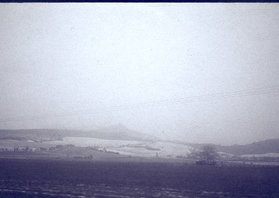 Vintage black-and-white landscape showing snow-covered hills under overcast skies. Distant farmland and sparse trees suggest ...