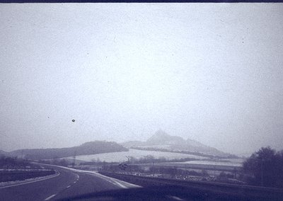 Vintage black-and-white roadside scene with misty mountains in background, likely mid-20th century. Curved road flanked by sp...