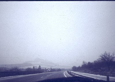 Vintage black-and-white roadside scene showing a two-lane highway with faint snow remnants. Distant, mist-covered mountain ra...