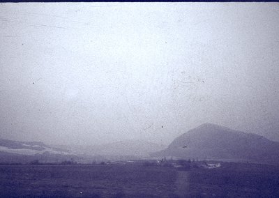Vintage sepia-toned coastal landscape with low-lying hills and sparse vegetation. Distant structures suggest a small settleme...