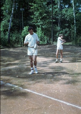 Two individuals in 1970s-style athletic wear—white shirts, shorts, and sneakers—play tennis on a worn outdoor court surrounde...