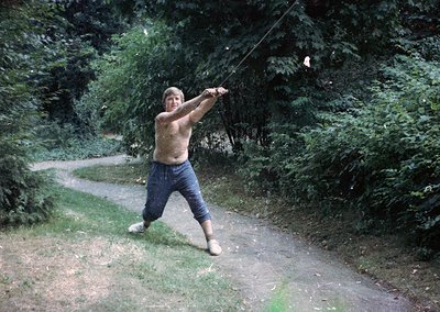 Shirtless man mid-action swinging a long wooden pole in a rural, wooded path, 1970s. Casual attire includes rolled-up trouser...