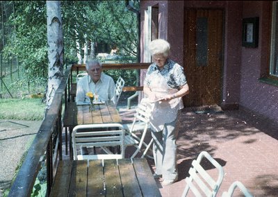 Vintage outdoor scene featuring two elderly individuals on a wooden balcony patio. Man seated at a table with a drink, woman ...