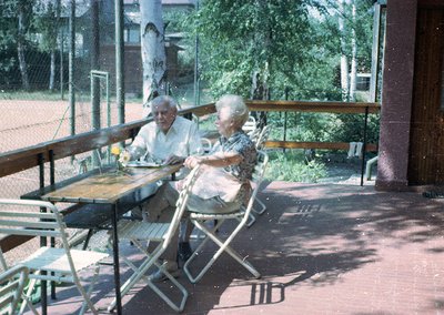 Two elderly individuals seated at an outdoor café table, likely mid-20th century (). Man in light-colored shirt, woman in pat...