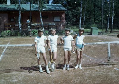 Four young boys in 1970s-style tennis attire pose on a clay court, holding vintage rackets. Wooden cabins and birch trees fra...