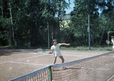 Vintage tennis player mid-forehand shot on a clay court, 1960s-70s. White short-sleeve shirt and shorts with a wooden racket....