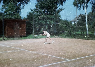 Vintage tennis court scene with player mid-swing, likely 1960s-70s. Clay surface, wooden changing hut, and lush greenery in b...