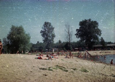 Vintage beach scene with sandy shore and shallow water, likely 1960s–1970s. Groups of people sunbathe, swim, and wade in natu...