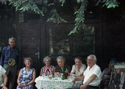 Vintage group portrait in rustic wooden cabin, likely 1970s. Six adults seated at a table with floral tablecloth, posing for ...