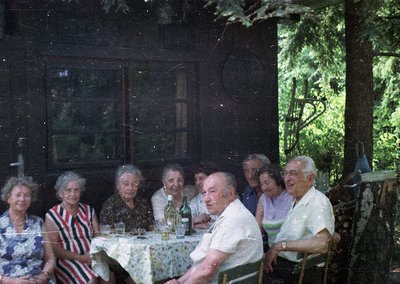 Vintage group portrait of nine elderly individuals seated at a rustic wooden table in a shaded outdoor setting, likely a gard...