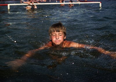 Young boy swimming in a pool, likely mid-1970s–1980s outdoor setting. Note the vintage swim trunks, shallow water, and blurre...
