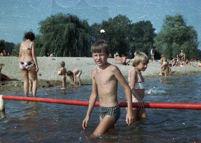 Vintage beach scene with children playing in shallow water, 1960s-1970s. Central boy in striped swim trunks stands near red p...