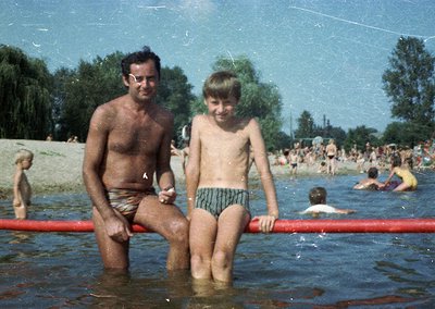 Vintage beach scene featuring a man and young boy in shallow water, likely mid-20th century. The man wears striped swim trunk...