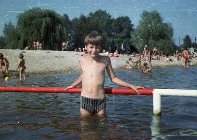 Shallow lakefront scene with a boy in striped swim trunks standing in water near a red pipe barrier. Lush green trees and gra...