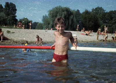 Vintage beach scene featuring a young boy in red swim trunks holding a surfboard, wading in shallow water. Surrounding him, o...
