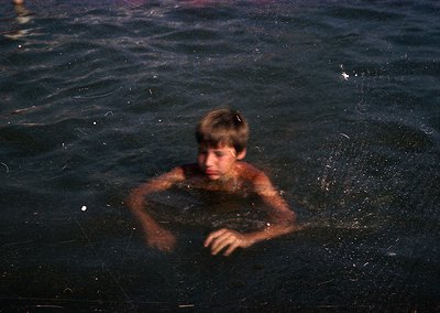 Young boy swimming in dark, shallow water, arms extended forward. Monochromatic, high-contrast tone suggests vintage or artis...