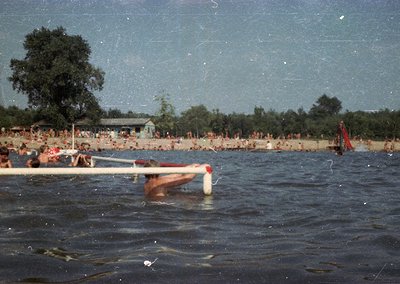 Vintage beach scene with crowded shore and waterfront. A lifeguard tower and wooden pier frame the foreground, while swimmers...