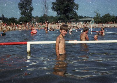 Vintage pool scene with young boy standing near a red-and-white goalpost in shallow water, surrounded by crowded swimmers. Li...