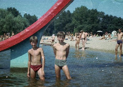 Two boys in wet swim trunks stand in shallow water under a red metal archway, likely part of a beach or pool entrance. Crowds...