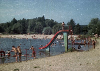 Vintage water park scene with red/blue slide and wooden platform, crowded with swimmers in 1960s-70s swimwear. Lush forested ...