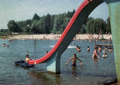 Vintage poolside scene featuring a red curved slide in a shallow pool, surrounded by swimmers in 1960s-70s swimwear. Lush gre...