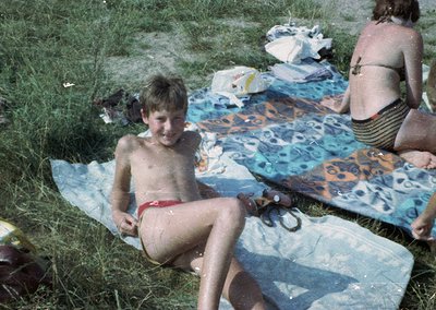 Vintage beach scene featuring a young boy in red swim trunks sitting on a patterned beach towel, surrounded by wet towels and...