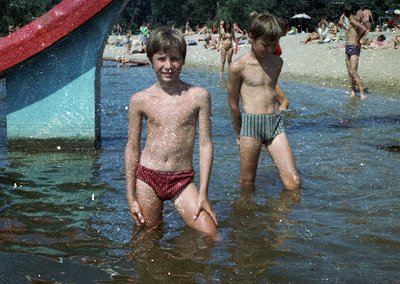 Two boys in red-striped swim trunks play in shallow water at a crowded beach, likely 1970s. Vibrant umbrellas and sunbathers ...