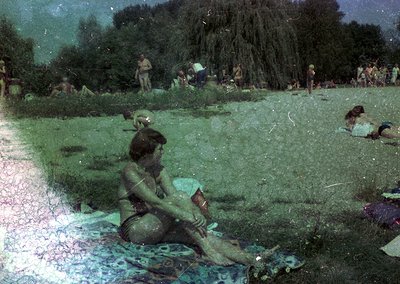 Vintage photo of a woman in a bikini reading a book on a patterned blanket in a grassy park. Mid-20th century summer scene wi...