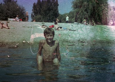 Vintage photo of a young boy playing in a shallow, algae-covered water body, likely a lake or river. Mid-20th century clothin...