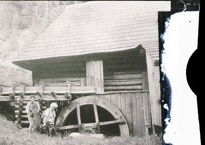 Vintage black-and-white photo of a rustic wooden waterwheel beside a log cabin, likely rural Europe, early-to-mid 20th centur...
