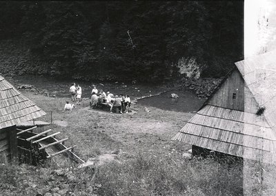 Black-and-white rural scene featuring a group of men in 1940s-50s attire gathering by a shallow stream, likely for water coll...