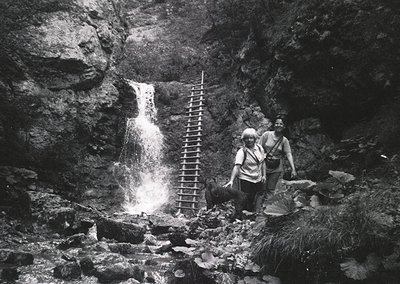 Mid-20th century black-and-white photo of two hikers beside a cascading waterfall in rugged alpine terrain. Ladder and rocky ...