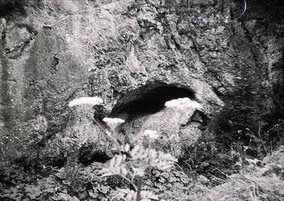 Black-and-white close-up of a cave entrance in rugged terrain, framed by jagged rock formations. Minimal vegetation at base s...