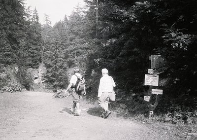 Two hikers in vintage outdoor gear stand on a dirt forest trail beside a wooden signpost marking "Jungfraujoch" and "Schiltho...