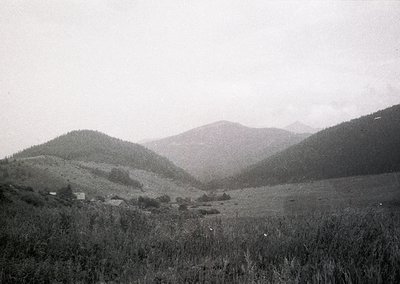Vintage black-and-white landscape of rolling hills and sparse vegetation, likely European alpine terrain. Small, scattered st...