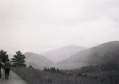 Mid-20th century black-and-white hike on winding mountain trail with misty valley views. Two hikers in casual 1950s-60s attir...