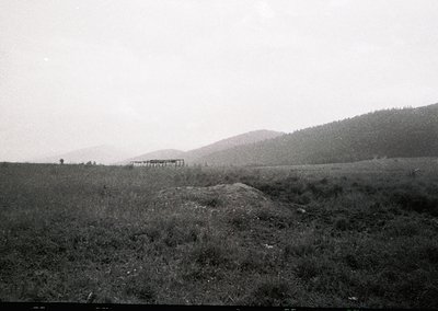Vintage black-and-white rural landscape featuring a modest wooden structure on a slight hillside, surrounded by open fields a...