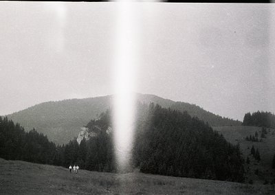 Vintage black-and-white mountain landscape with three hikers on a grassy ridge, framed by dense forest and rocky peaks. Sunli...
