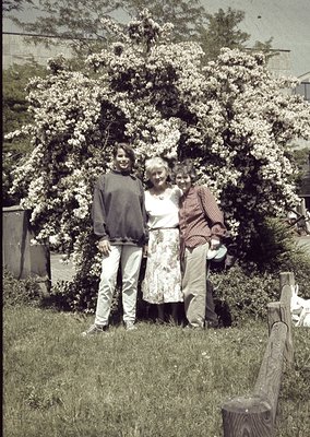 Three individuals pose in a lush garden under blooming trees, likely hawthorn, in a sepia-toned photo. The woman wears a flor...