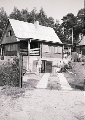 Mid-century alpine-style wooden chalet with gabled roof and brick chimney, set in a forested area. A man in shorts stands nea...