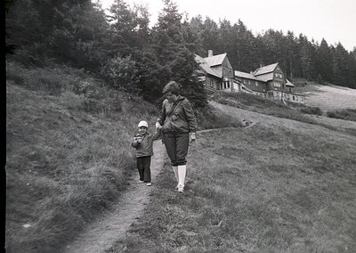 Mid-20th century black-and-white photo of an adult and child walking on a grassy hillside path, leading toward rustic wooden ...