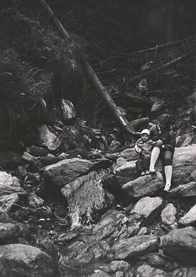 Mid-century black-and-white photo of a lone woman in a sleeveless dress and stockings navigating rocky terrain, likely a hiki...
