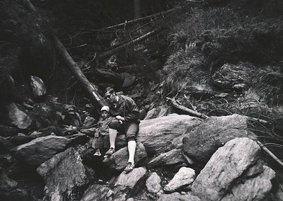 Black-and-white shot of a climber mid-ascent on a rugged rock face, secured by ropes and harness. The climber wears a cap, sh...