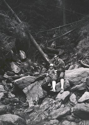 Two men in early 20th-century outdoor attire—wide-brimmed hats, long-sleeve shirts, and knee-high socks—rest on rugged rocks ...