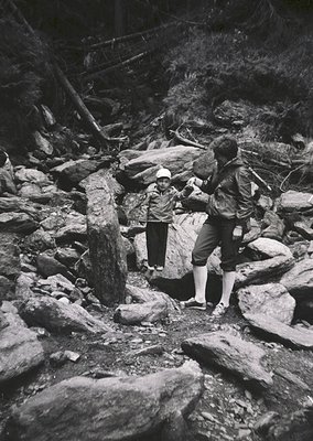 Mid-20th century black-and-white photo of two hikers navigating rocky alpine terrain. Adult assists child across uneven stone...