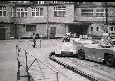 Vintage 1960s–70s courtyard scene featuring a Ford-based go-kart race with "McLaren" branding on a small kart. Spectators, in...