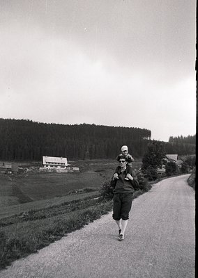A woman in mid-20th-century outdoorwear—wide-brimmed hat, long-sleeve shirt, and knee-length skirt—walks a child on a rural r...