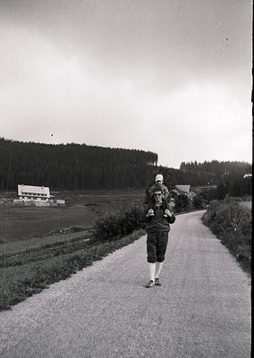 Mid-20th century rural roadside scene: Woman in 1950s-style dress and headscarf carries child on back, walking beside paved r...