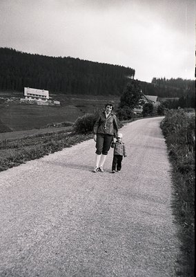 A woman and child walk a rural road flanked by dense forest and open fields, likely mid-20th century. The adult wears a long ...